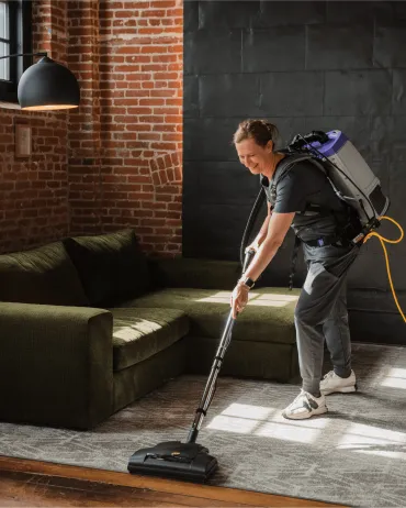 Merry Maids of Berks County cleaner vacuuming a living room rug with a backpack vacuum near a sectional sofa